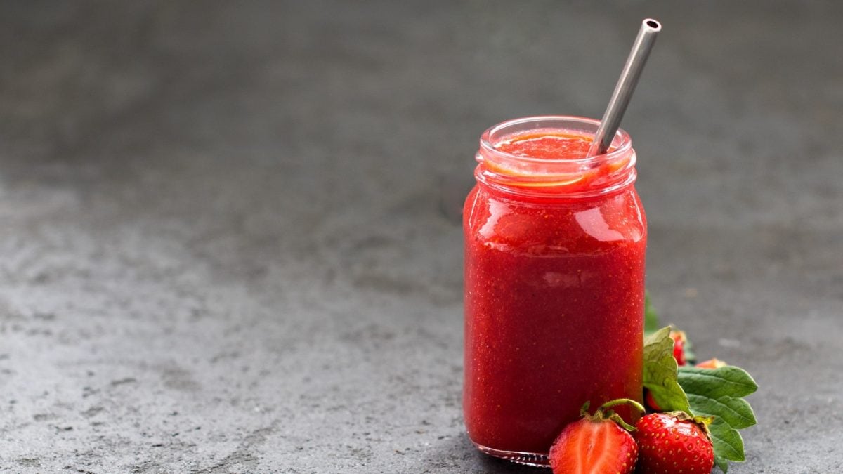 Ein Glas Erdbeerlimo mit Mango und Banane und einem Strohhalm aus Metall, daneben liegen ein paar frische Erdbeeren.