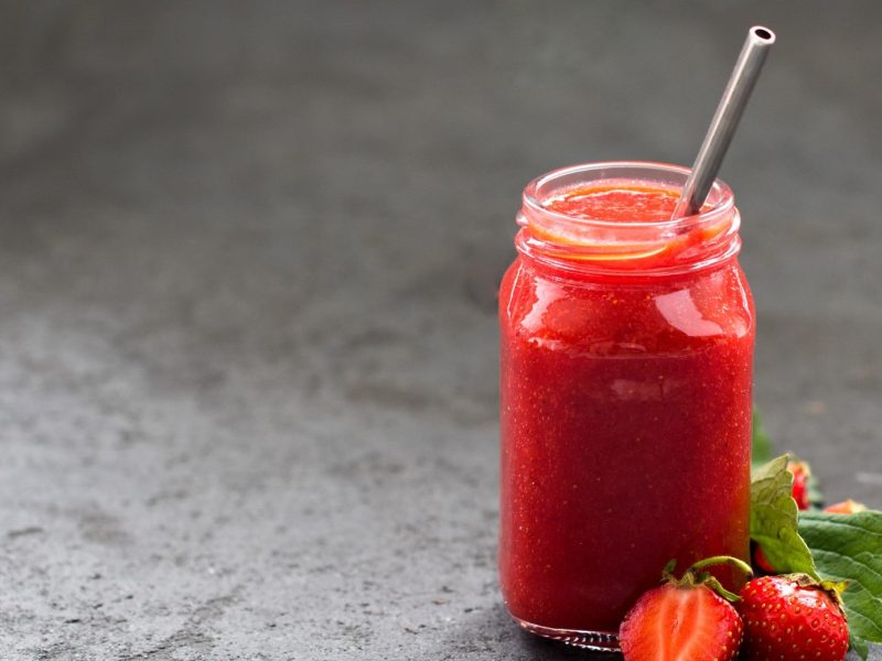 Ein Glas Erdbeerlimo mit Mango und Banane und einem Strohhalm aus Metall, daneben liegen ein paar frische Erdbeeren.
