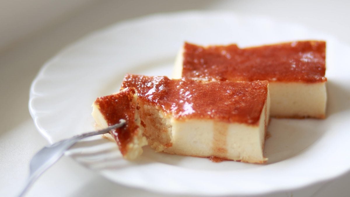 Galatopita griechischer Milchkuchen auf weißem Teller. Links Gabel, die einStück den Kuchen auslöst. Frontalansicht.
