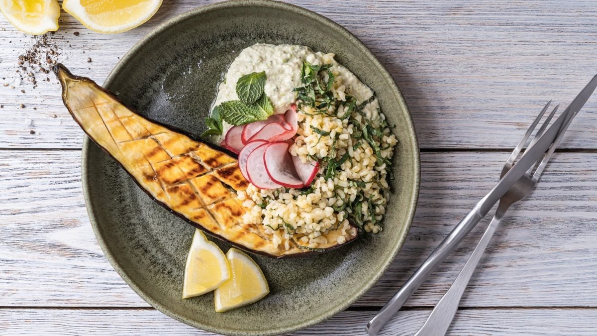 Ein Teller gebackene Augerbine mit Bulgur-Spinat-Salat, Radieschen, Minze und Zitronenspalten in der Draufsicht, daneben Besteck.