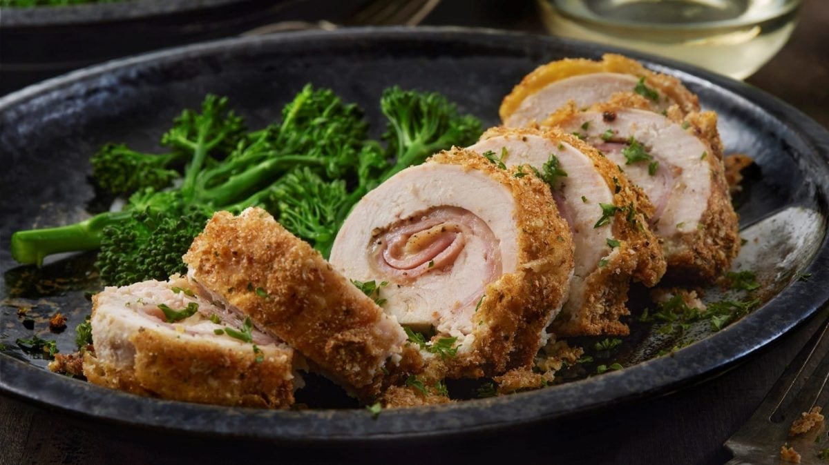 Gerolltes Cordon Bleu auf einem dunklen Teller neben Brocolini