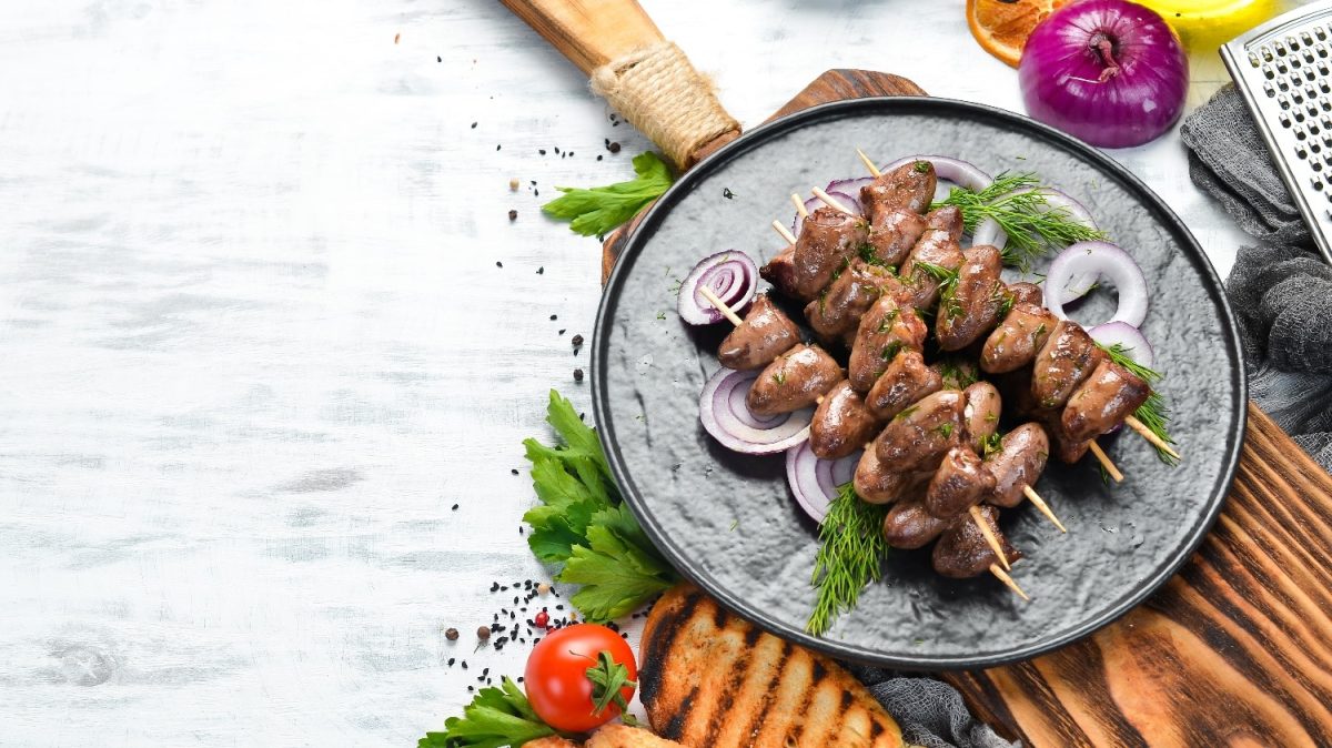 Ein grauer Teller mit den gegrillten Hühnerherzen auf einem Holzbrett. Drum herum einiges an Gemüse und Kräutern.