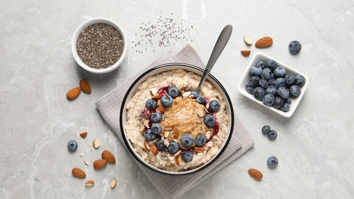 Eine Schale mit dem Mandel-Porridge auf einem hellgrauen Tisch. Um die Schale herum sind Mandeln, Blaubeeren und Chiasamen.