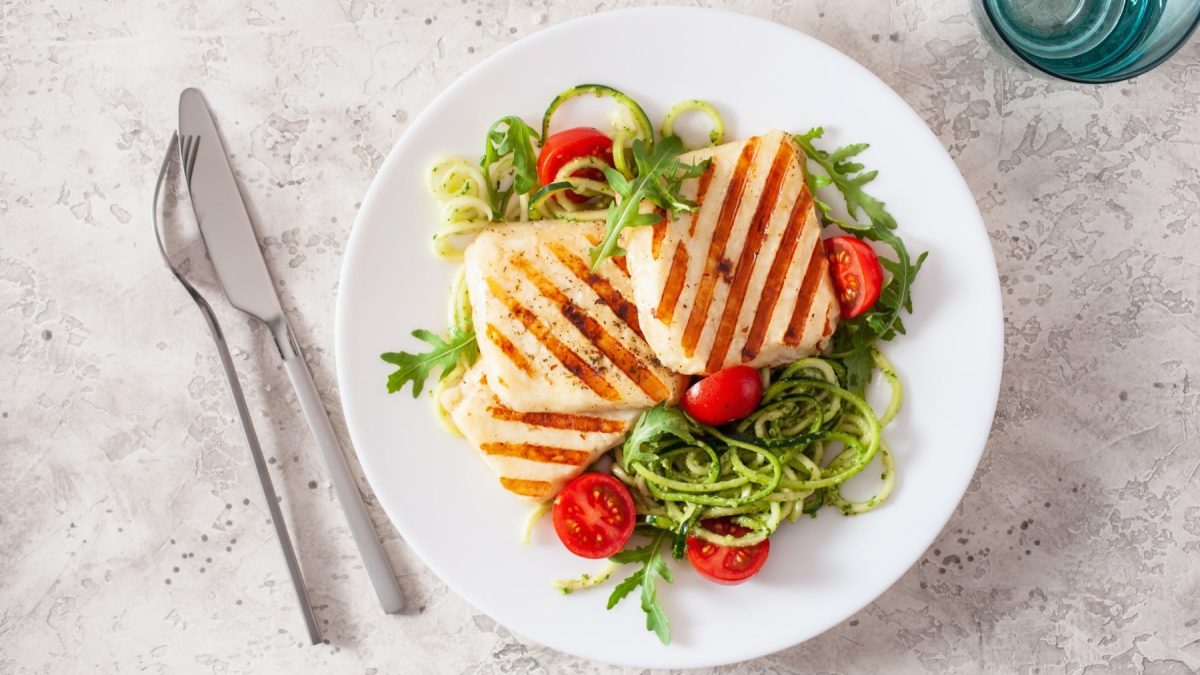 Halloumi auf Zucchinsalat mit grünem Pesto auf einem weißen Teller. Daneben liegt Besteck.