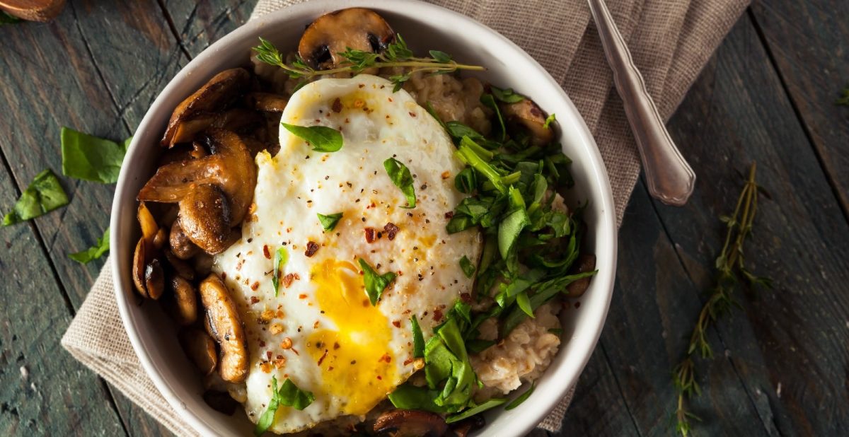 Eine Schüssel herzhafter Porridge mit Spiegelei, Spinat und braunen Champignons auf einem beigen Küchentuch.