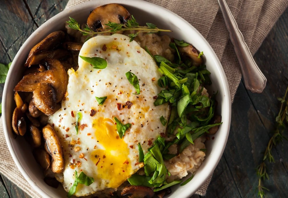 Eine Schüssel herzhafter Porridge mit Spiegelei, Spinat und braunen Champignons auf einem beigen Küchentuch.