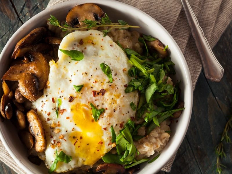 Eine Schüssel herzhafter Porridge mit Spiegelei, Spinat und braunen Champignons auf einem beigen Küchentuch.