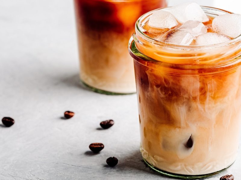 Ein Glas Iced Vanilla Latte mit Eiswürfeln, daneben liegen Kaffeebohnen.