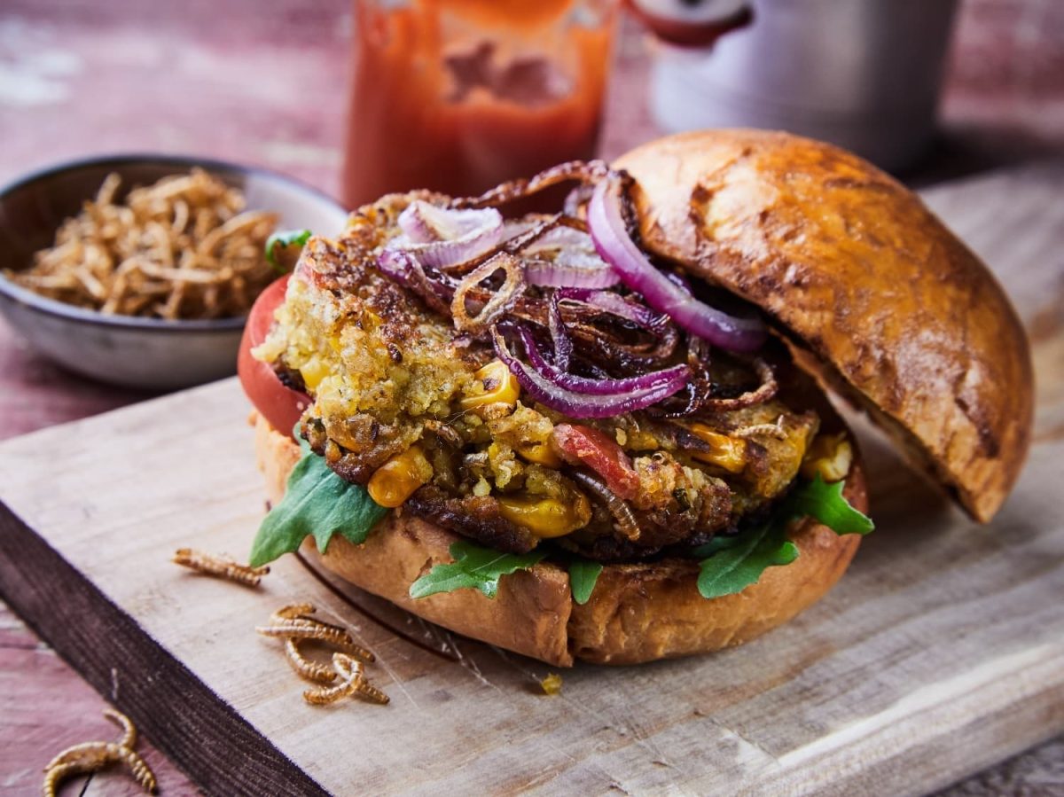 Ein Burger mit Insekten auf einem Holzschneidebrett. Im Hintergund eine Schale mit mehr Würmern und Ketchup im Glas.