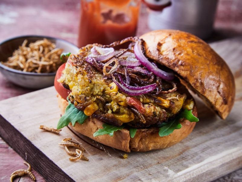 Ein Burger mit Insekten auf einem Holzschneidebrett. Im Hintergund eine Schale mit mehr Würmern und Ketchup im Glas.