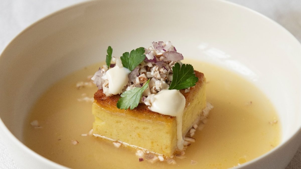 Eine weiße Schale mit goldgelber Brühe und einem viereckigen Kartoffelpudding in der Mitte des Tellers. Auf dem Kartoffelpudding sind Champignons, Creme Double und etwas Petersilie.