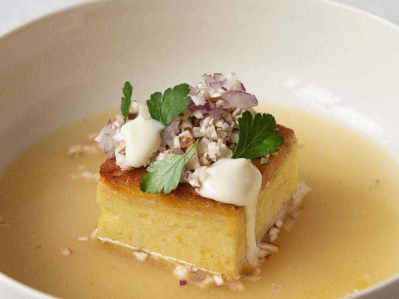 Eine weiße Schale mit goldgelber Brühe und einem viereckigen Kartoffelpudding in der Mitte des Tellers. Auf dem Kartoffelpudding sind Champignons, Creme Double und etwas Petersilie.