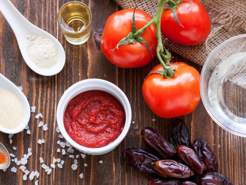 Ketchup ohne Zucker selber machen mit Datteln. Tomaten, Datteln, Salz sowie Schalen und weiße Löffel mit den Zutaten auf Holztisch. Aufnahme aus der Vogelperspektive.