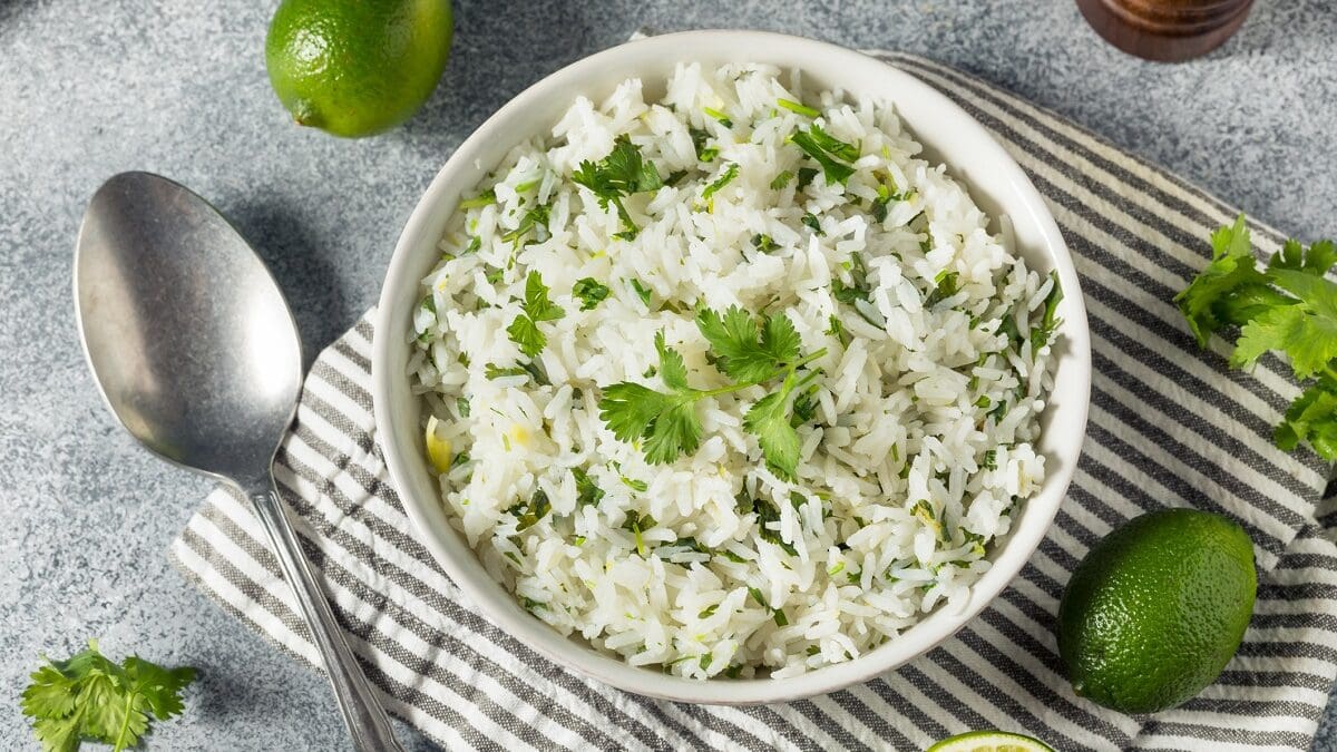 Eine Schale Koriander-Limetten-Reis steht auf einem Tisch. Daneben liegen ein Löffel sowie Limetten.
