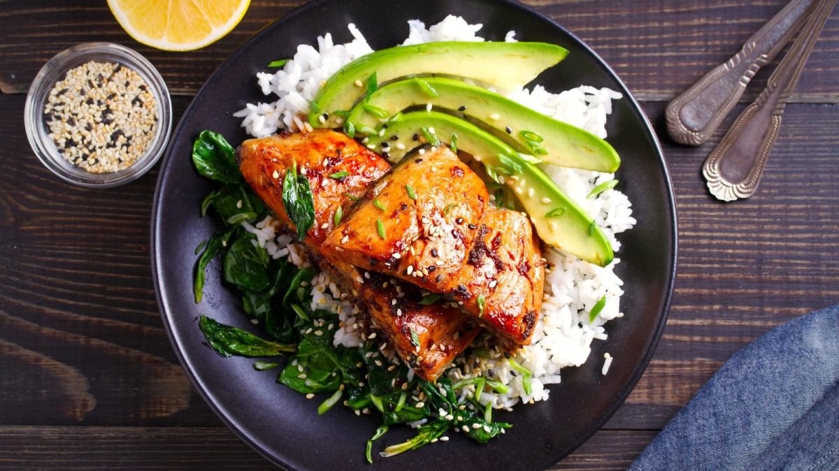 Lachs mit Honig-Soja-Glasur,, Avocado, Spinat und Reis auf einem schwarzen Teller auf einem holzfarbenen Tisch