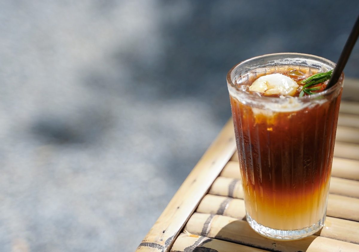 Ein Glas mit dem Litschi-Eistee und einem schwarzen Strohhalm auf einem Holztisch und Betonuntergrund.