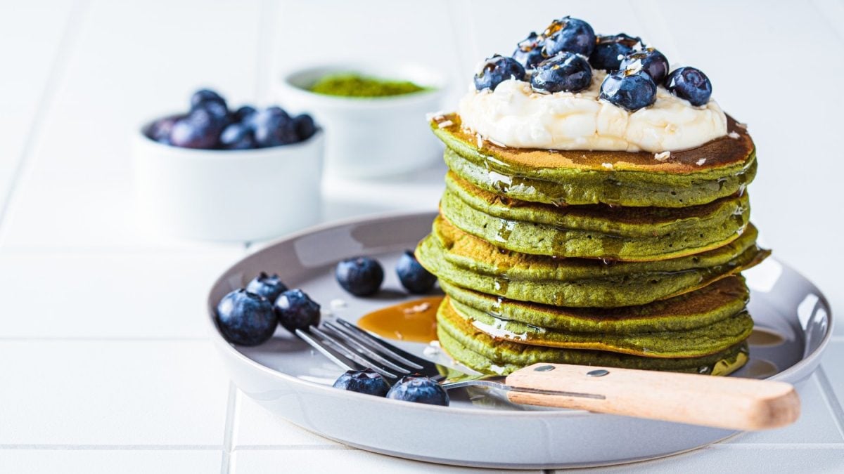 Matcha-Pancakes auf einem grauen Teller. Im Hintergrund zwei Schüsseln mit Heidelbeeren und Matchpulver.