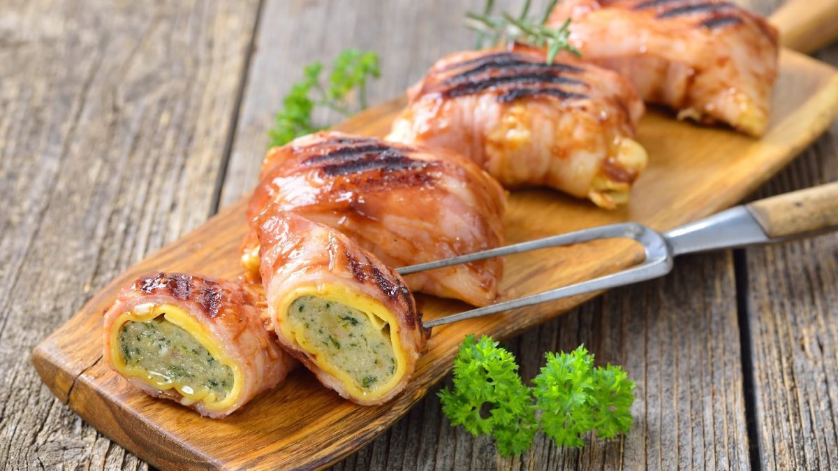 Fünf Maultaschen mit Speck bardiert und gebraten auf einem langen Holzbrett, welches wiederum auf einem Holztisch steht.