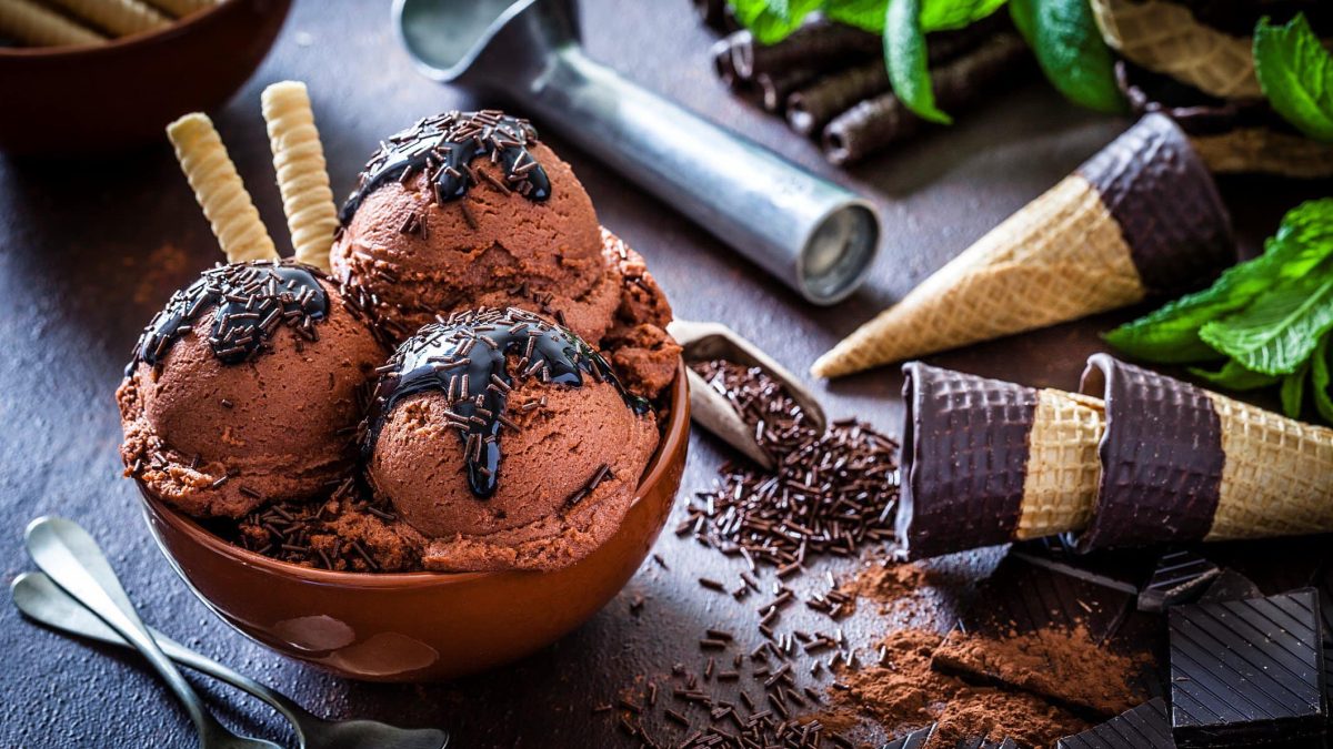 Selbst gemachtes Milchmädchen Schokoladeneis ohne Eismaschine in Schale mit Schokoladensauce und -streuseln auf dunklem Untergrund. Daneben Eiswaffeln mit Schokoladenrand. Eislöffel am Bildrand sichtbar. Draufsicht.