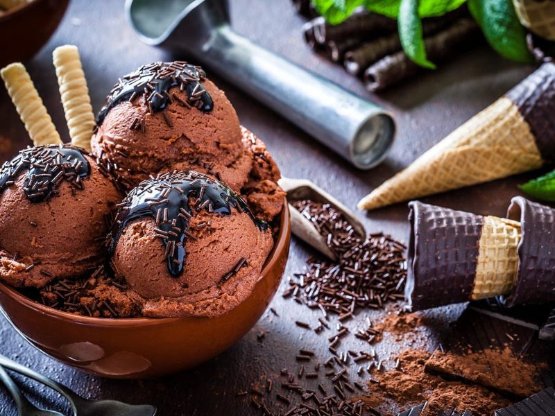 Selbst gemachtes Milchmädchen Schokoladeneis ohne Eismaschine in Schale mit Schokoladensauce und -streuseln auf dunklem Untergrund. Daneben Eiswaffeln mit Schokoladenrand. Eislöffel am Bildrand sichtbar. Draufsicht.