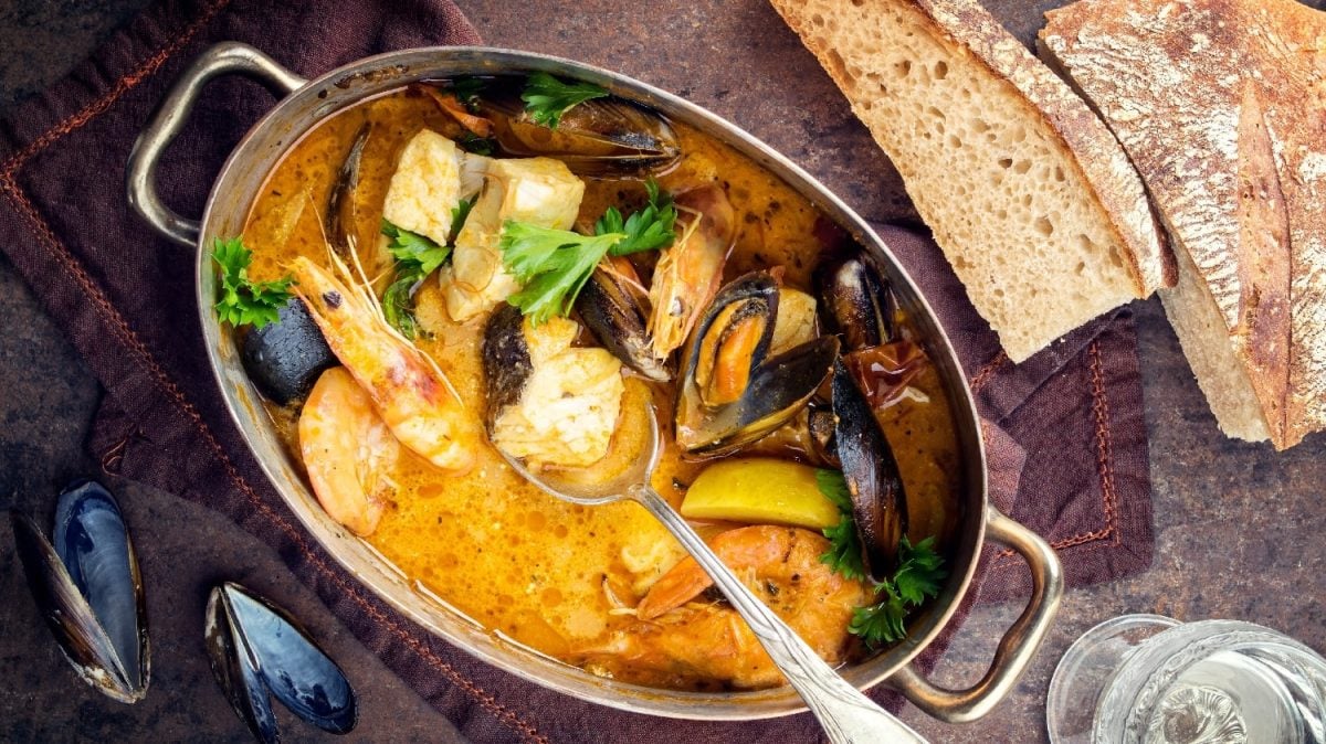 Ein brasilianischer Fischeintopf in einem silberne ovaler Topf. Darunter einige Miesmuscheln und darüber etwas Brot auf einem dunkelbraunen Untergrund.