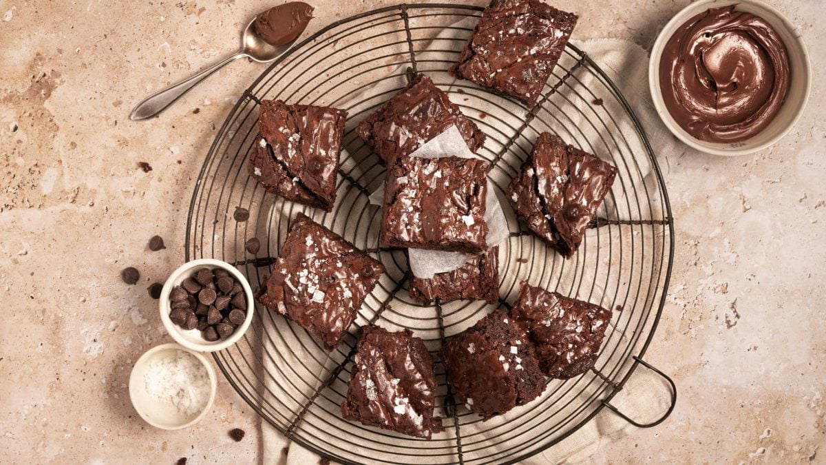 Nutella-Brownies auf Kuchengitter auf hellbraunem Untergrund. Daneben Schälchen mit Meersalz, Schokodrops und Nutella. Oben Teelöffel mit Nutella. Aufnahme aus der Vogelperspektive.