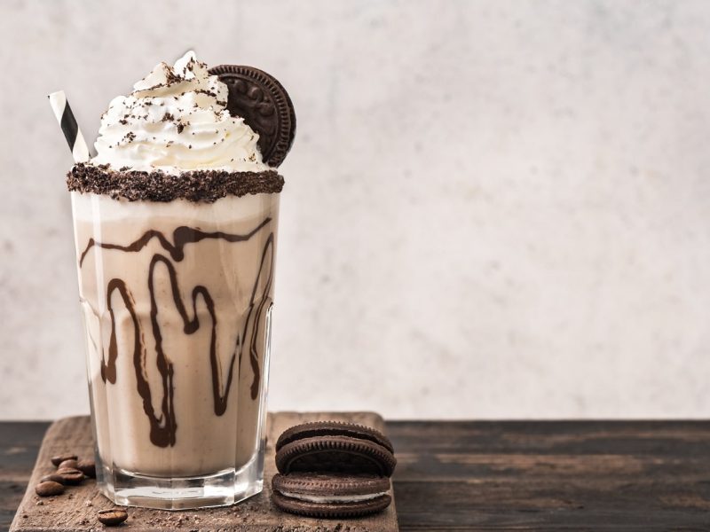 Ein Glas Schokokeks-Milchshake mit Strohhalm, Sahnehaube und Keks, daneben liegen zwei Kekse.