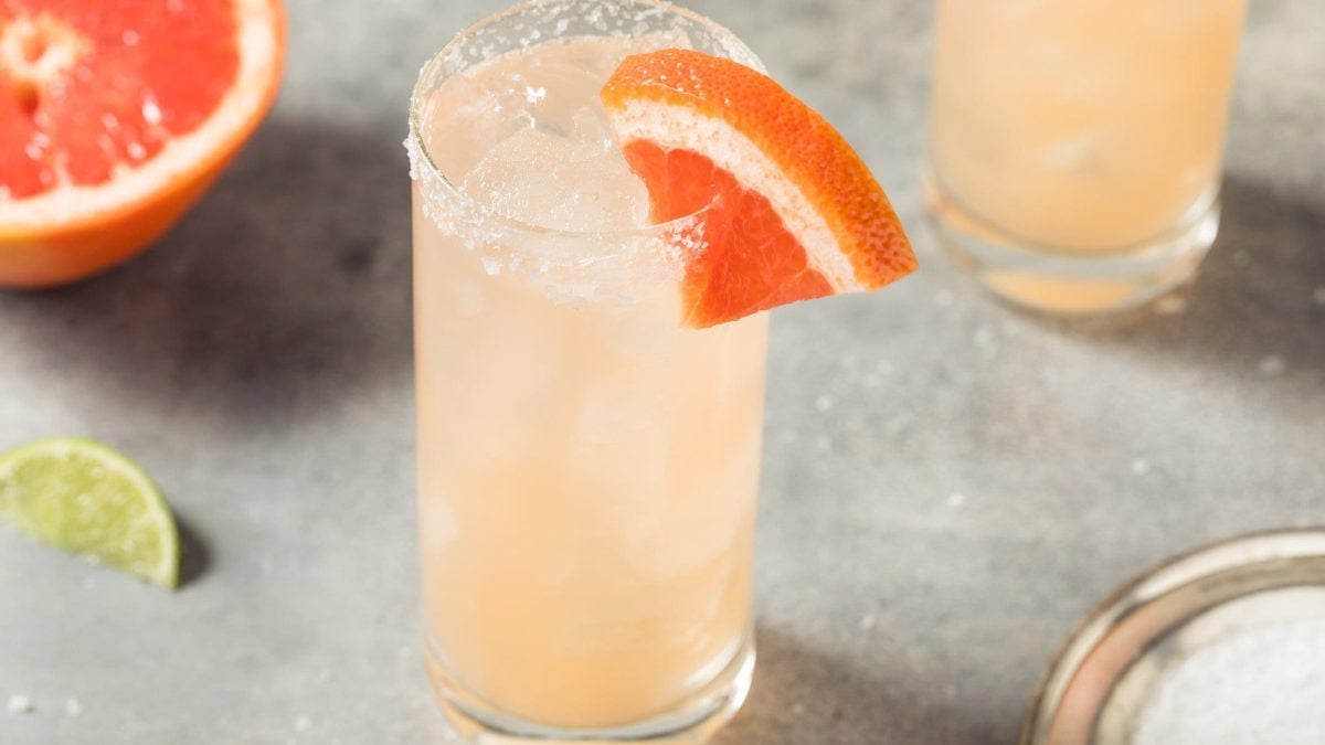 Paloma mit Blutorange Cocktail mit Tequila in Highball-Glas auf grauem Untergrund. Im Bild Blutorange und Limette. Oben ein weiteres Glas angedeutet. Draufsicht.