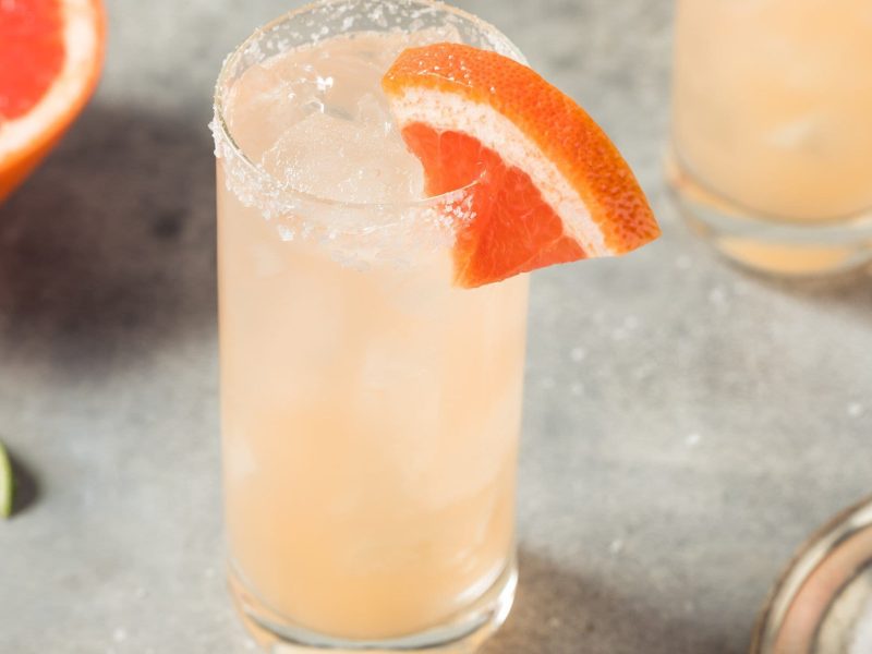 Paloma mit Blutorange Cocktail mit Tequila in Highball-Glas auf grauem Untergrund. Im Bild Blutorange und Limette. Oben ein weiteres Glas angedeutet. Draufsicht.