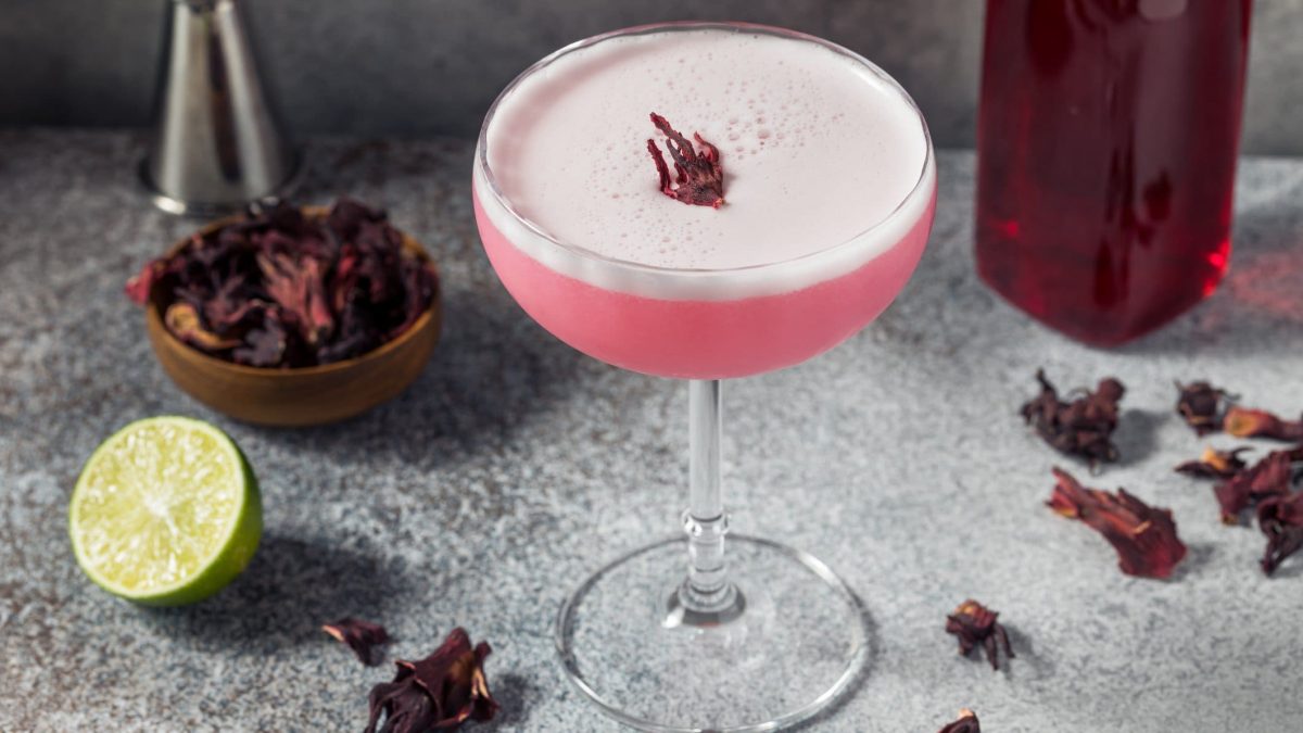 Pink Vodka Sour mit Hibiskus infused Vodka auf grauem Tisch. Daneben Hibiskus-Blüten, Limette und Bar-Maß. Recht eine Flasche mit Hibiskus-Sirup. Frontalansicht.