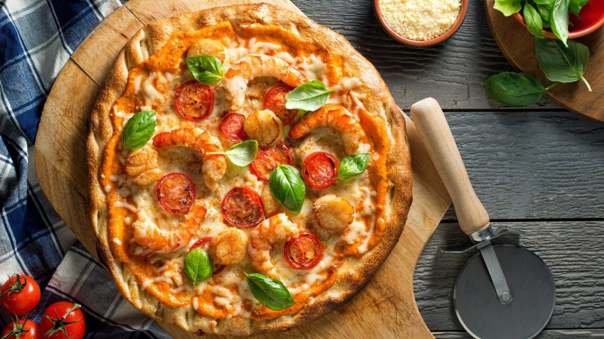 Pizza Gamberetti auf einem Holzbrett. Daneben liegen verschiedene Zutaten.