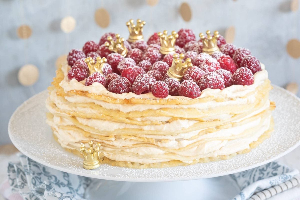 Eine Pfannkuchentorte mit Karamellquark und Himbeeren, dazwischen kleine goldene Krönchen, auf einem Tortenständer. Das Rezept stammt aus dem Kochbuch "Es war einmal".