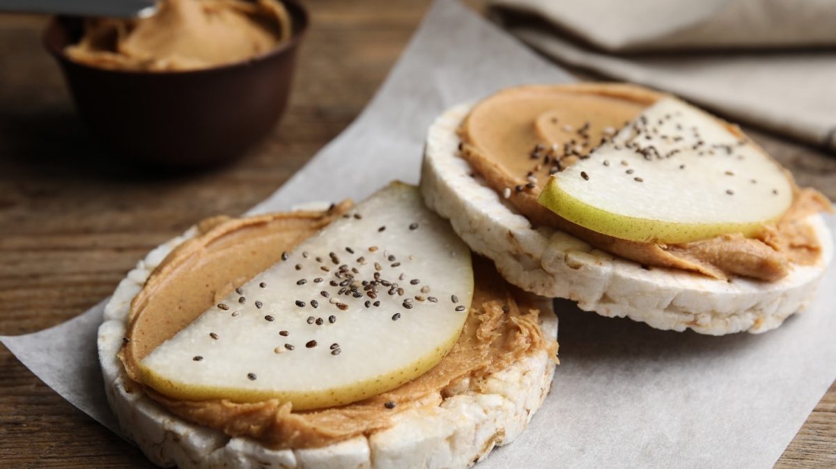 Zwei Reiswaffeln mit hellbrauner Creme auf Pergamentpapier, welches wiederum auf einem Holztisch liegt. Außerdem ist eine Birnenspalte und Chiasamen auf den Reiswaffeln.