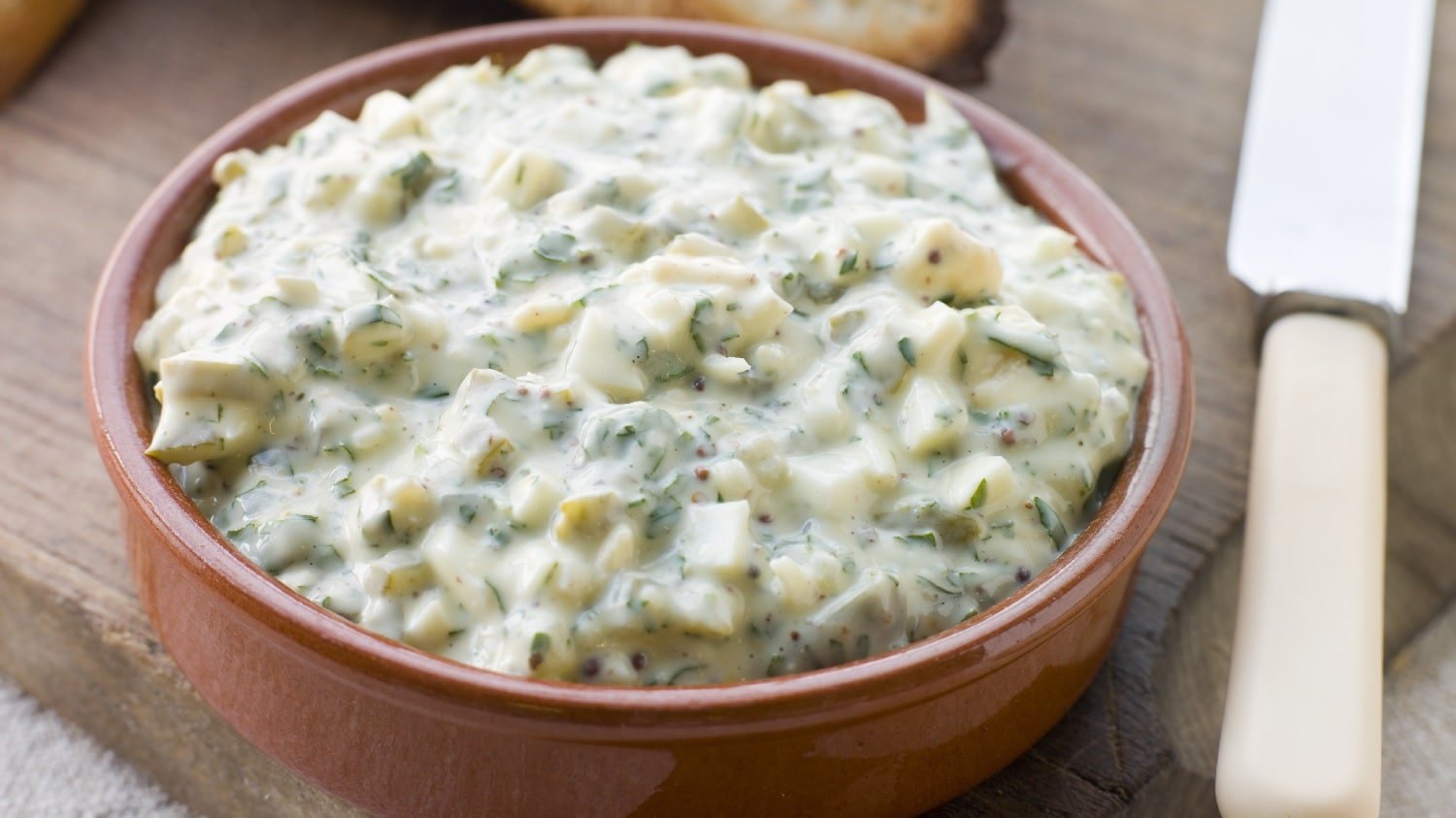 Eine braune Schale mit der Sauce Gribiche auf einem Holztisch. Im Vordergrund ein Messer und im Hintergrund ein paar geröstete Scheiben Weißbrot.