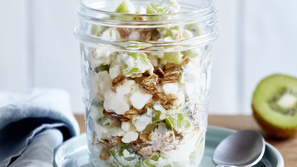 Schicht-Flocken aus dem Glas mit Kiwi stehen auf einer Untertasse. Daneben ein Löffel und eine aufgeschnittene Kiwi.