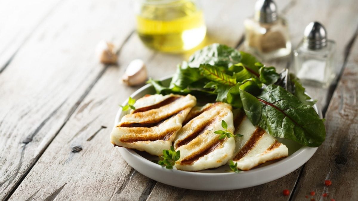 Ein Teller mit ein paar Scheiben selbst gemachtem veganem Halloumi und grünem Salat, dahinter unscharf Öl, Gewürzstreuer und Knoblauchzehen.