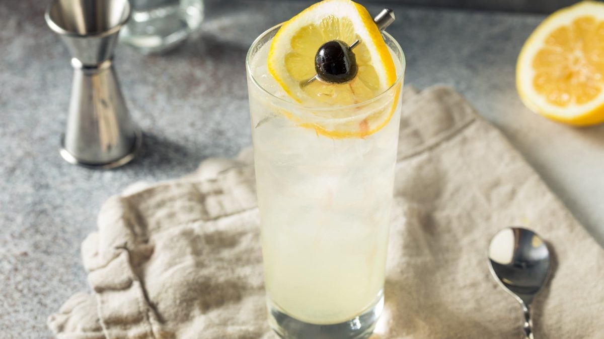 Tom Collins Gin-Cocktail auf beigem Tuch auf grauem Tisch mit Zitronenscheibe und Kirsche auf Metallspieß. Daneben Barmaß. Im Hintergrund eine halbe Zitrone. Frontalansicht.