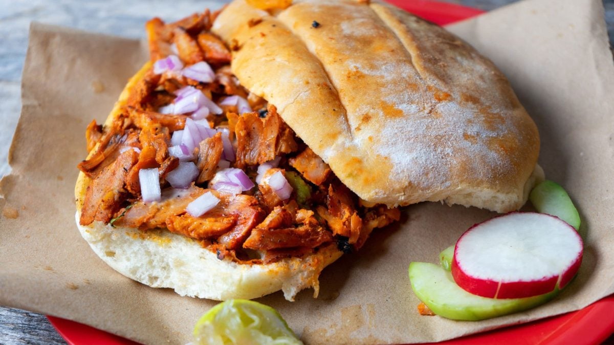 Tortas al Pastor auf braunem Papier auf rotem Teller auf blauem Untergrund. Daneben eine Scheibe Radieschen und Limette. Draufsicht.