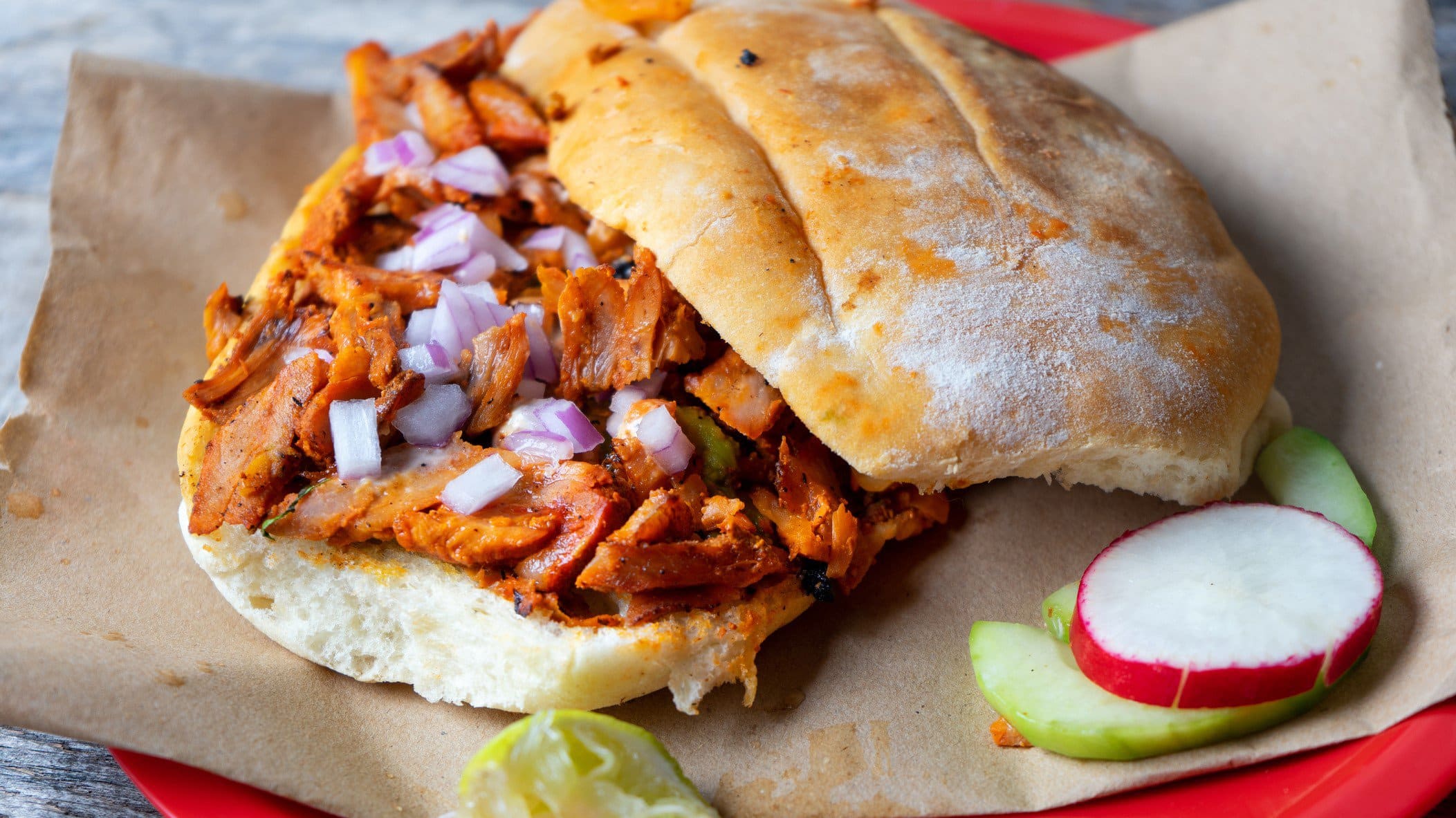 Tortas al Pastor auf braunem Papier auf rotem Teller auf blauem Untergrund. Daneben eine Scheibe Radieschen und Limette. Draufsicht.