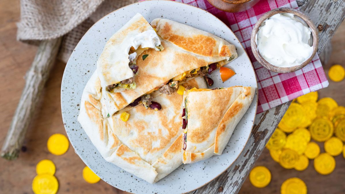 Tortilla-Crunchwraps mit Hackfleisch auf einem Teller in der Draufsicht, daneben eine Schale mit weißem Dip und ein paar Goldtaler.