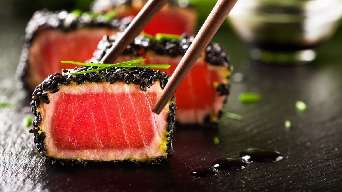Tunatataki mit schwarzem Sesam und Miso-Creme auf schwarzer Granitplatte. Im Hintergrund Sauce in Glasschüssel. Zwei Stäbchen von oben ins Bild ragend. Frontalansicht.