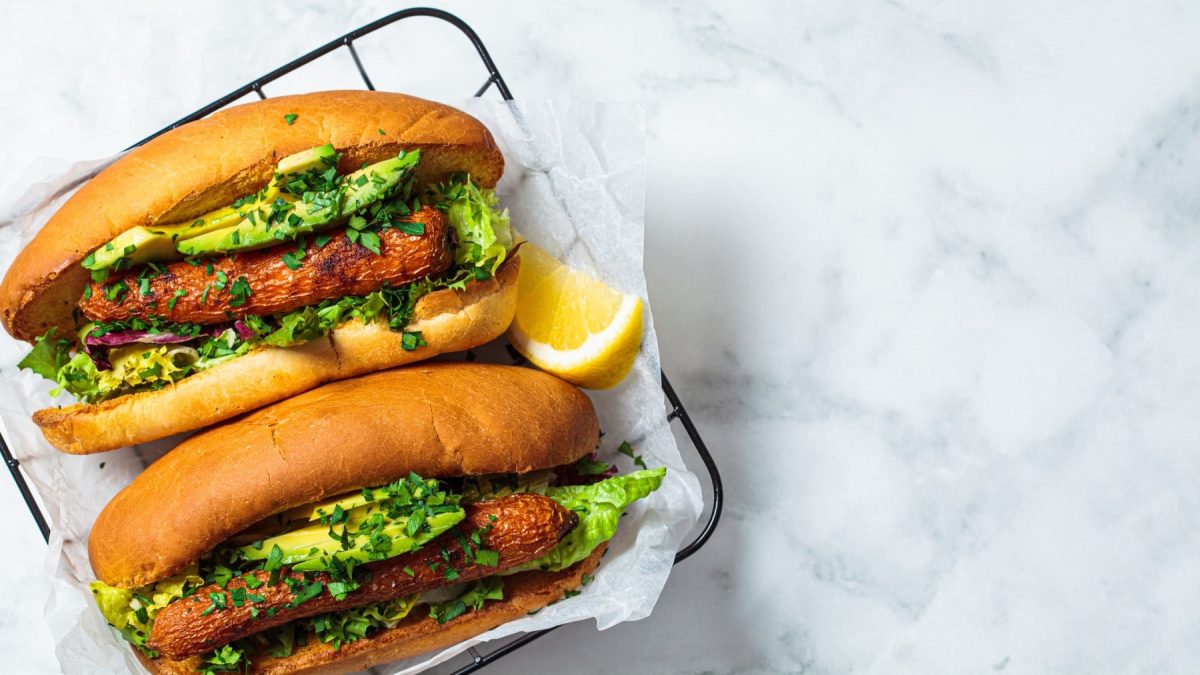 Vegane Hotdogs auf Backpapier auf einem schwarzen Gitter. Daneben liegt eine Zitrone.