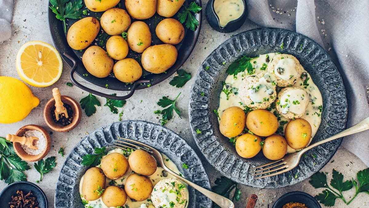 Zwei Teller vegane Königsberger Klopse mit Pellkartoffeln in der Draufsicht, außerdem ein Topf Kartoffeln und Schalen mit Gewürzen.