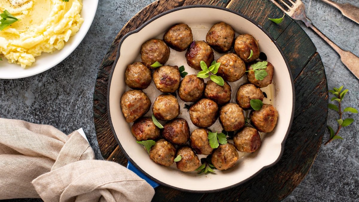 Eine Pfanne vegane Köttbullar mit Kräutern bestreut auf einem runden Brett in der Draufsicht, daneben eine Schale Kartoffelpüree, ein Geschirrtuch und eine Gabel.