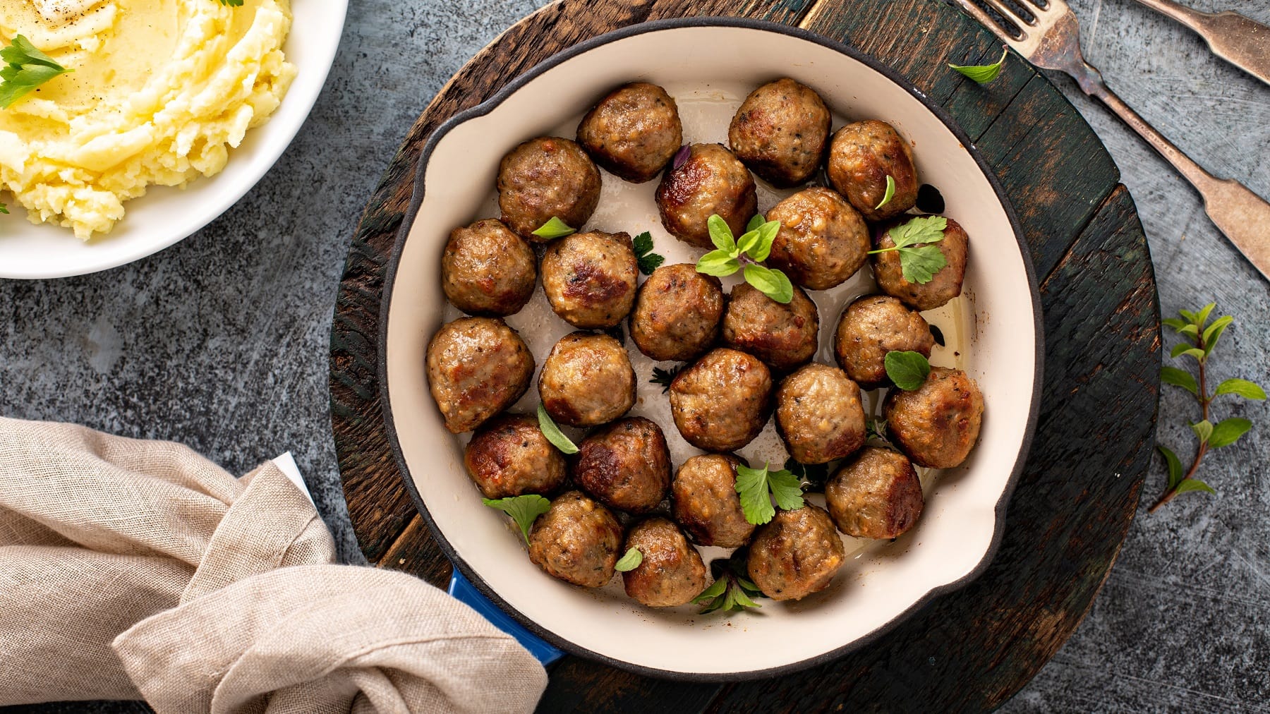Eine Pfanne vegane Köttbullar mit Kräutern bestreut auf einem runden Brett in der Draufsicht, daneben eine Schale Kartoffelpüree, ein Geschirrtuch und eine Gabel.