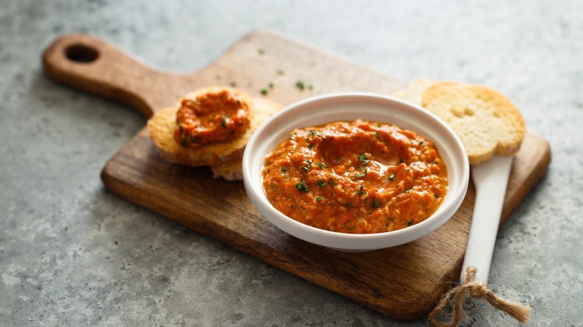 Eine Schale veganer Tomate-Walnuss-Aufstrich steht auf einem Brett, auf dem auch zwei bestrichene Baguettescheiben liegen.