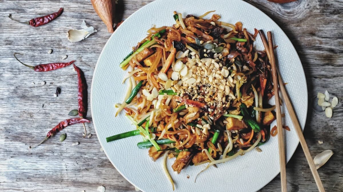 Ein Teller veganes Pad Thai mit Erdnüssen, Frühlingszwiebeln und Möhren, daneben Essstäbchen. Alles in der Draufsicht.