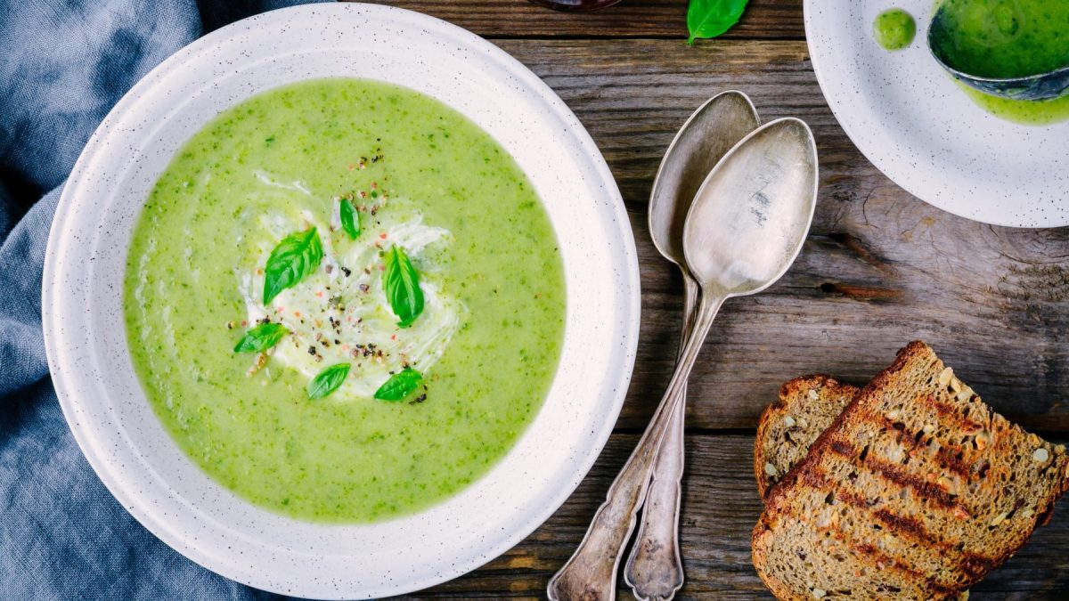 Zucchinicreme-Suppe mit Gorgonzola auf einem weißen Teller. Daneben zwei Scheiben Brot und zwei Löffel.