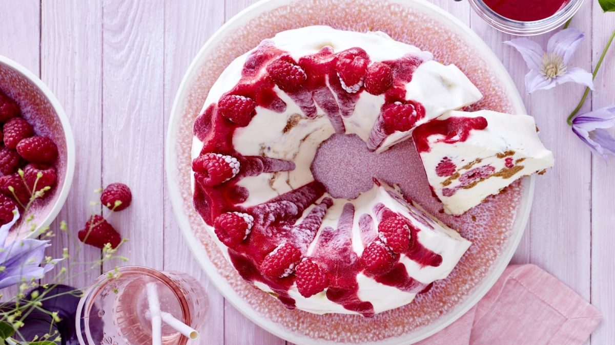 Auf einem Teller steht ein Cheesecake-Eis-Gugelhupf. Er ist mit Himbeeren belegt und mit Soße überzeugen.