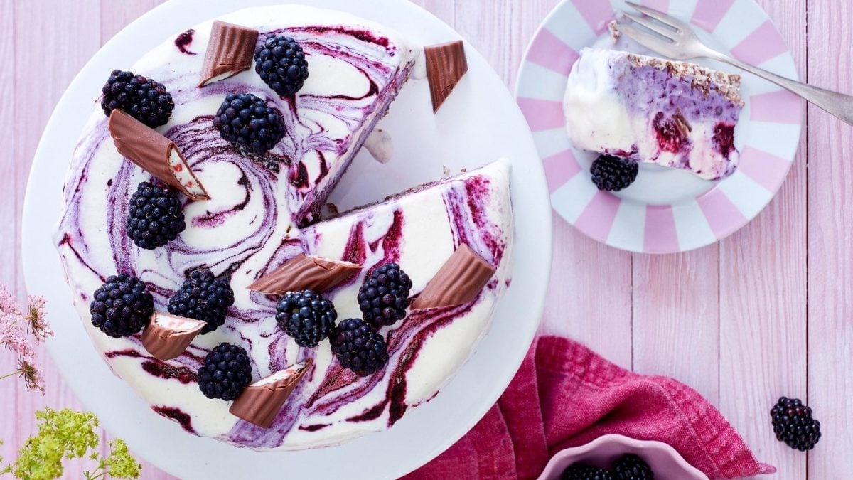 Eistorte mit Brombeeren und Joghurt, angeschnitten serviert auf einem hellen Teller, garniert mit frischen Brombeeren und Schokoladen-Stückchen auf einem hellen Tisch.